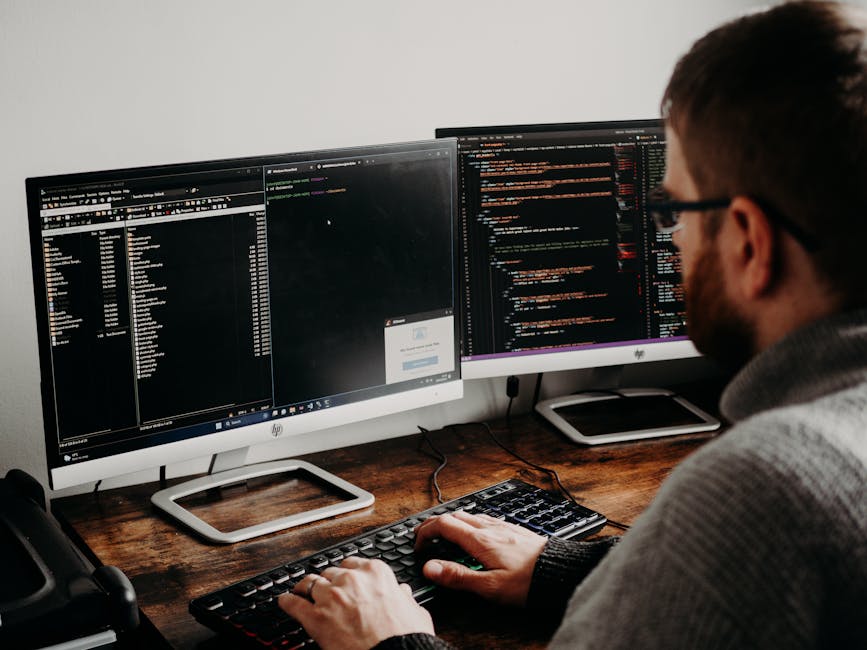 Software developer typing code on dual monitors at a wooden desk